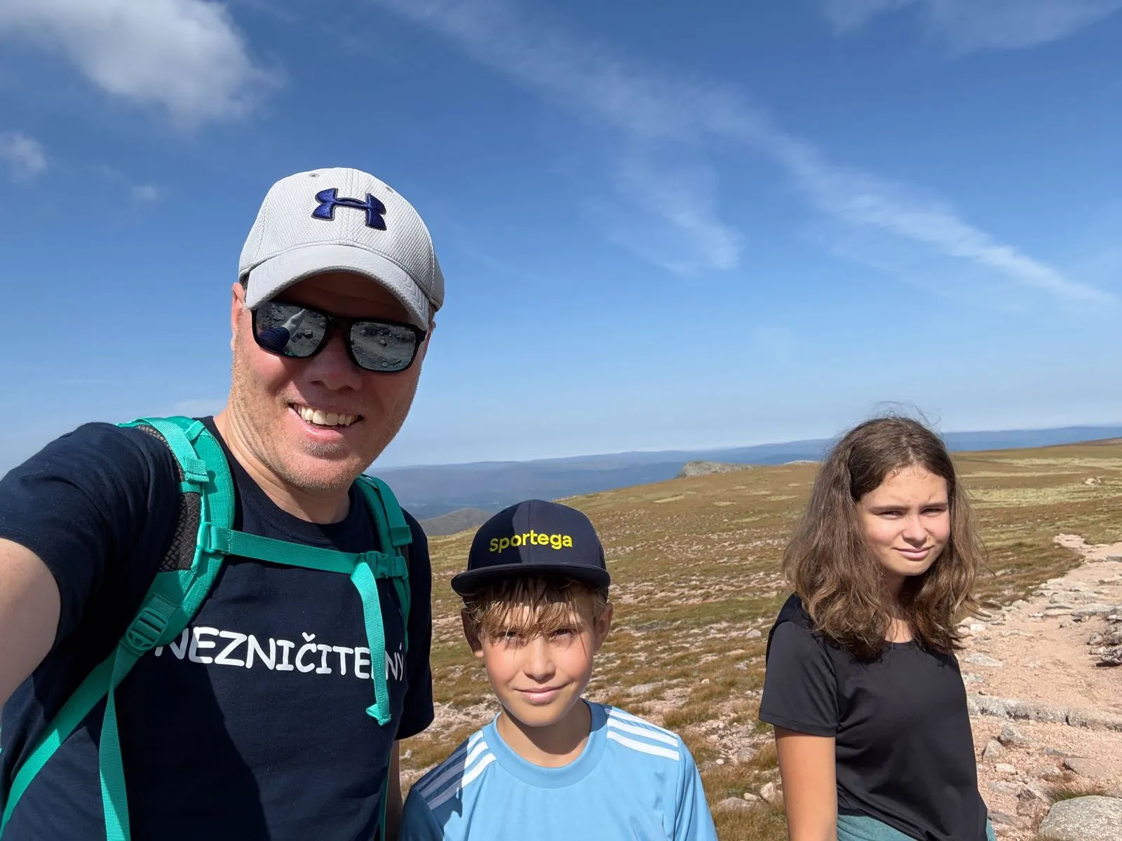 Skupina tří osob v horách, s panoramatickým výhledem na krajinu, v pozadí modrá obloha a zelené kopce.