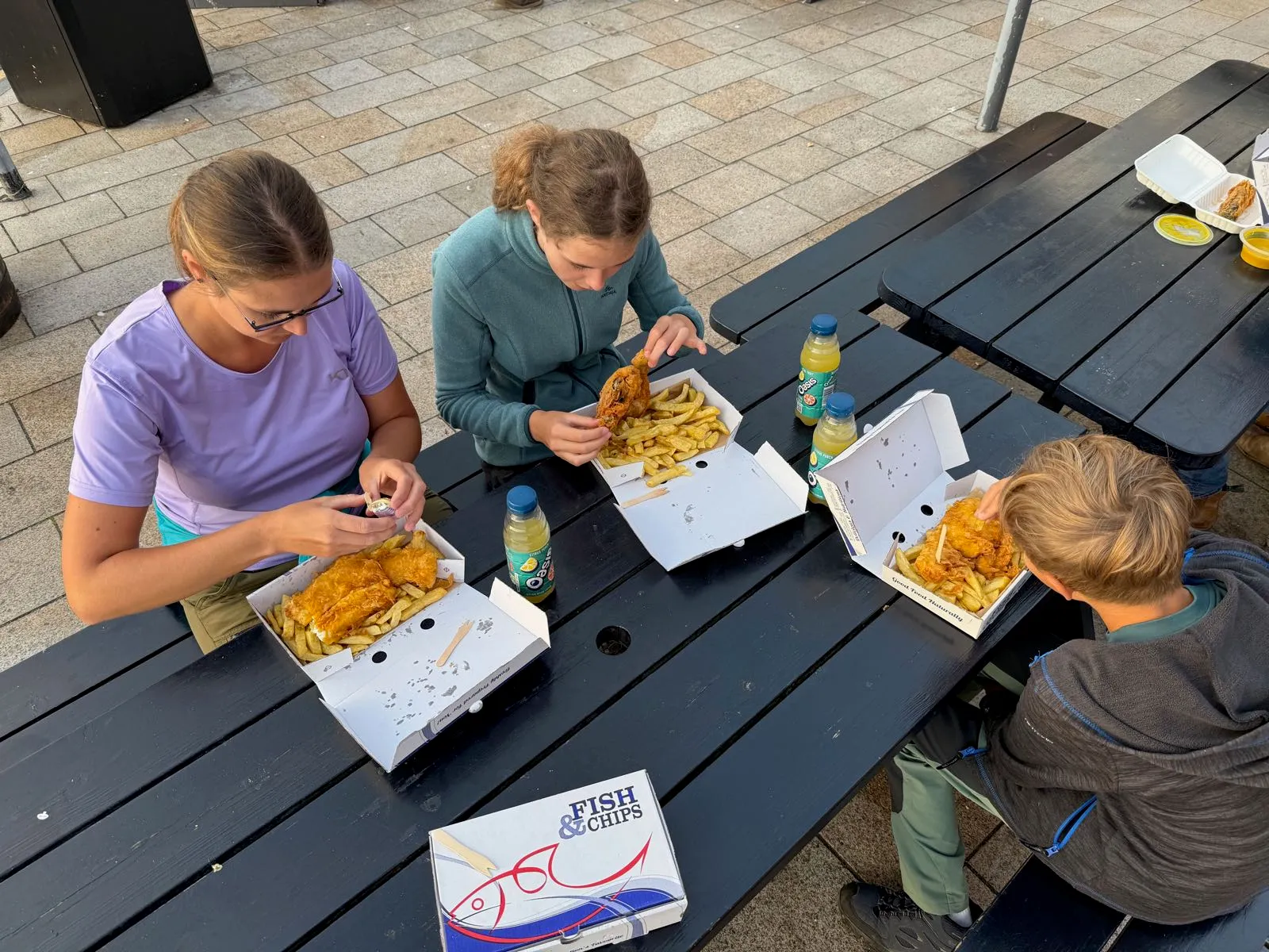 Rodina vychutnává fish and chips u stolů na venkovní terase. Na stole jsou otevřené krabičky s pokrmem a nápoji.