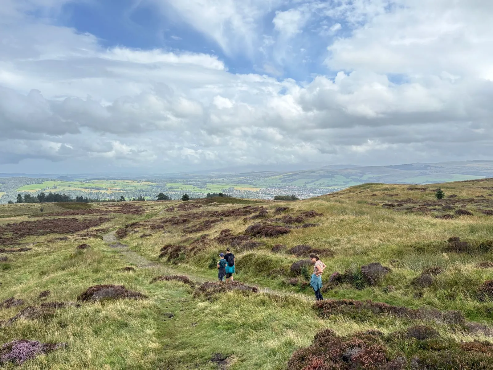 Two hikers walking on a path through green rolling hills under a cloudy sky in Scotland.