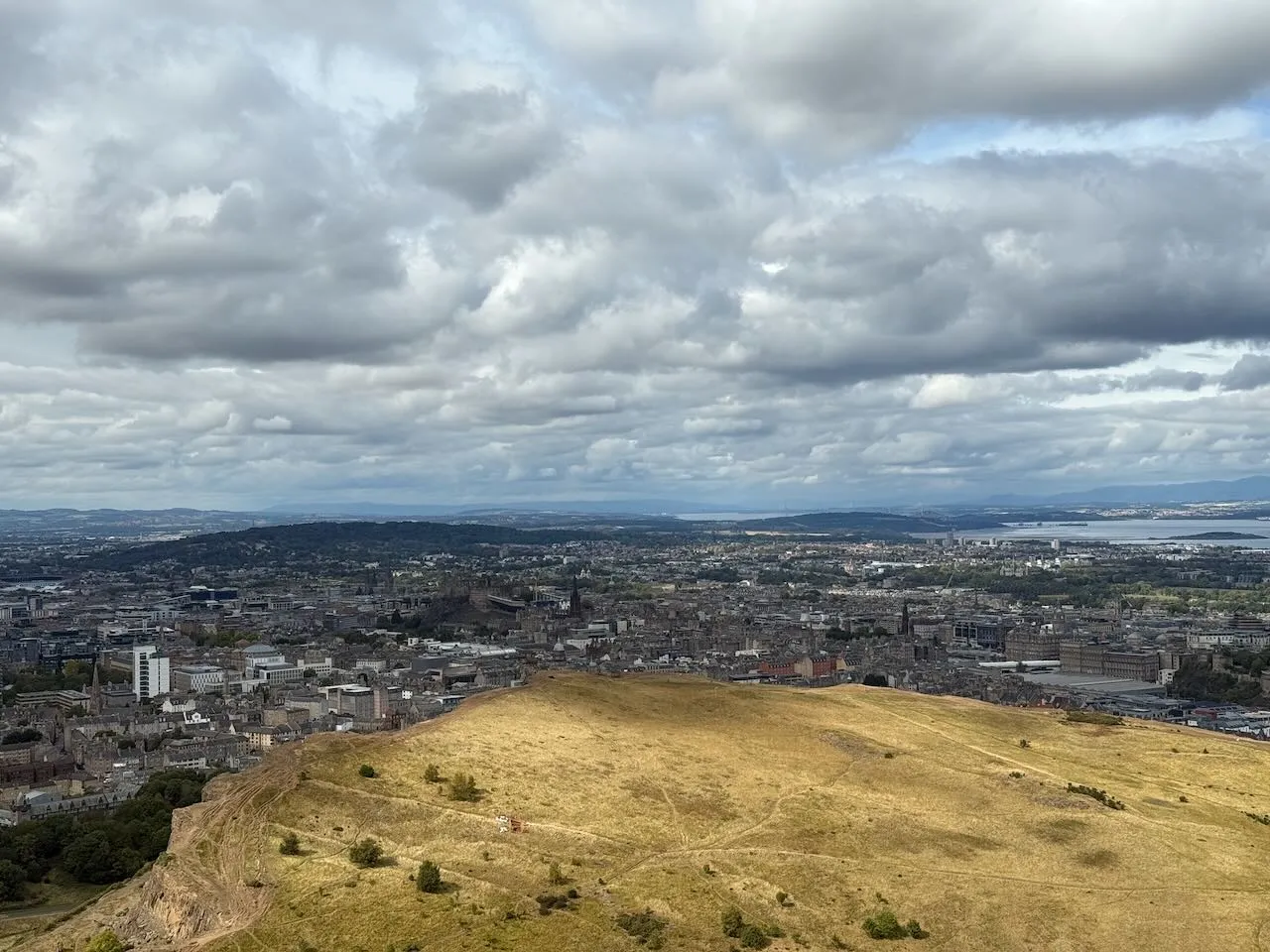 Pohled na Edinburgh z vrcholu Arthur’s Seat, s městskou krajinou a oblačným nebem v pozadí.