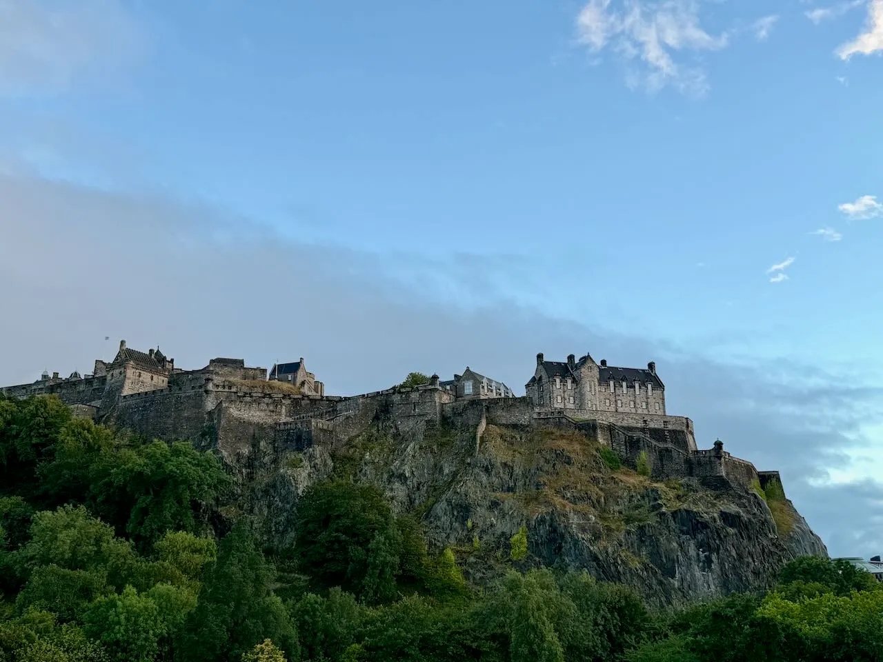 Obrázek Edinburghu s hradem na kopci, zasazený do modré oblohy s mraky.
